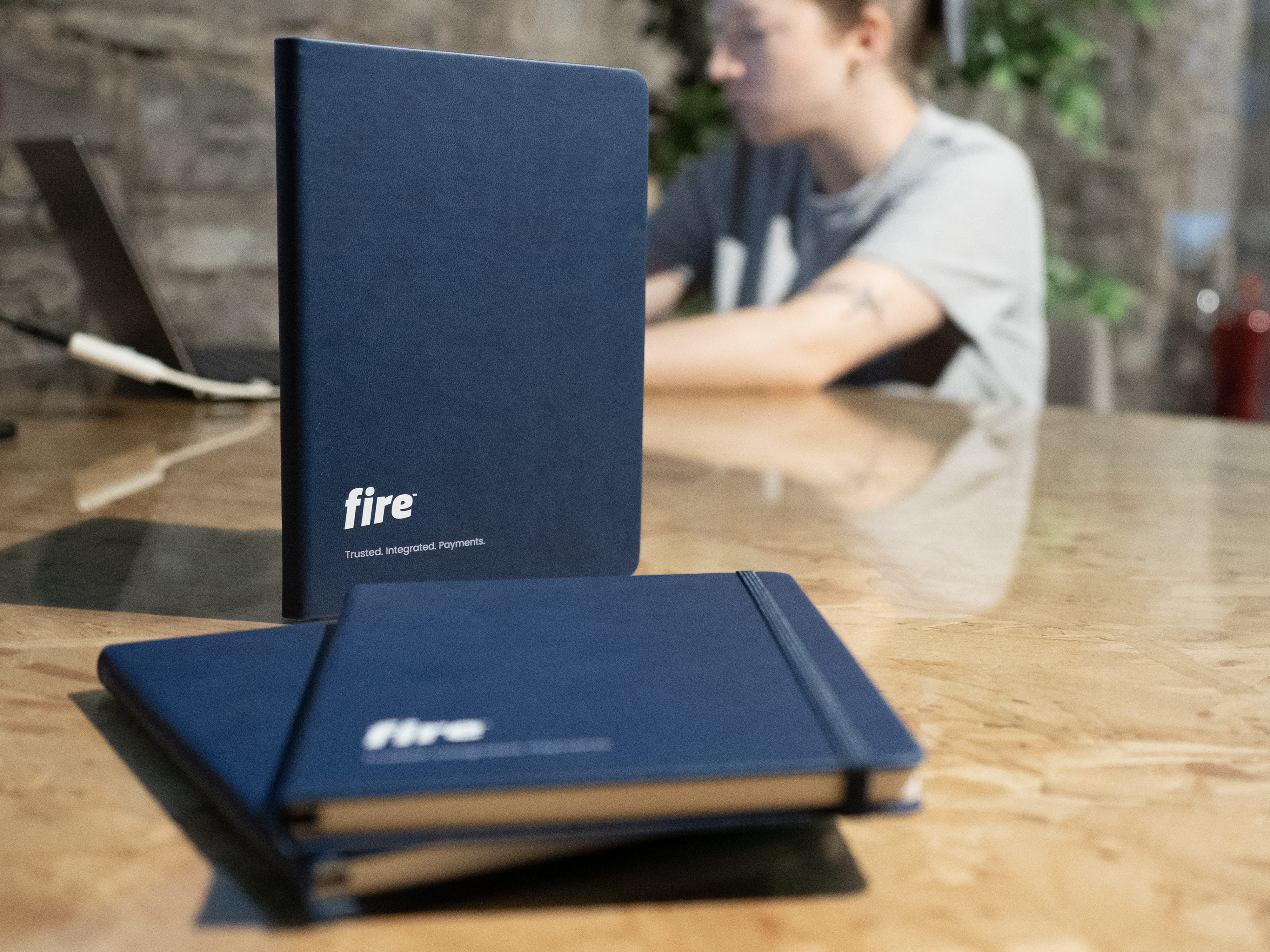 Dark blue branded notebooks with the Fire logo are placed on a wooden table, while a person sits in the background working on a laptop in a modern office setting.