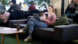 A woman sits on a grey couch in a modern, collaborative workspace. She is smiling while looking at her phone, with a laptop on her lap. She wears a pink jumper, jeans, and trainers. On the wooden coffee table in front of her is a Fire branded water bottle.