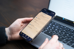 A person holding a smartphone displaying the Fire Business mobile application showing a €10,000 transfer from a main account, while working on a laptop. The screen displays transaction details, including currency conversion and account information.