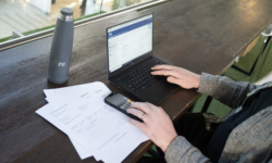 A person sits at a wooden table working on a laptop displaying the Fire web app. They are using a smartphone calculator while referencing printed invoices on the table. A grey Fire-branded water bottle is placed nearby.