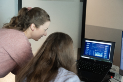Two women are looking at a laptop screen displaying a data dashboard with graphs, charts, and financial metrics. One woman is seated, while the other leans over, engaged in conversation.