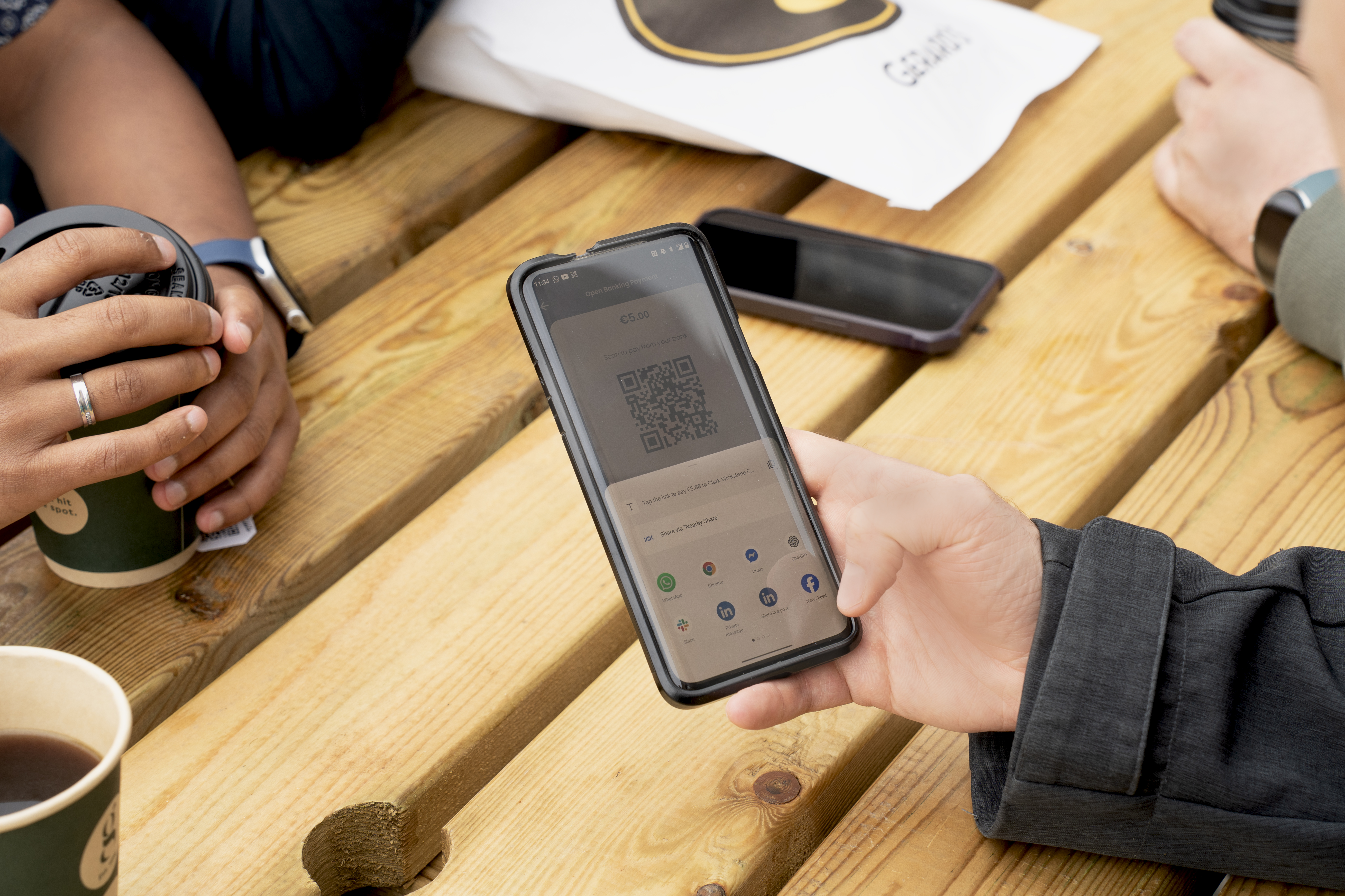Hand holding a phone showing Fire’s open banking portal, on a wooden table with coffees and people in the background.
